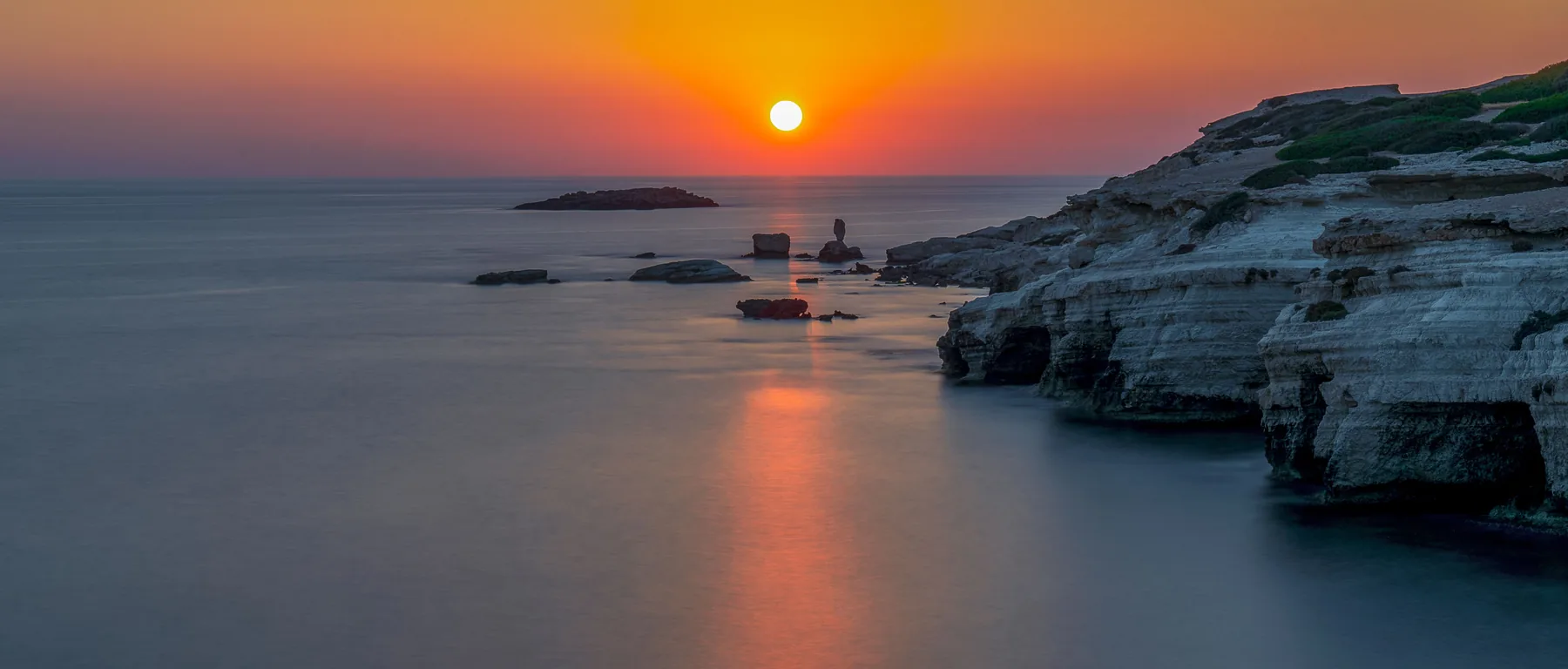 A pale limestone coastline and a small island on calm water as the sun sets over the horizon